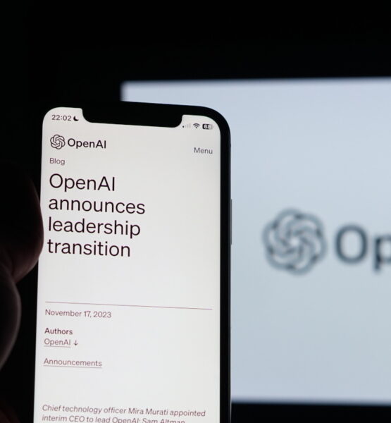 Ein Smartphone, das in der Hand gehalten wird, zeigt einen Blogbeitrag von OpenAI mit der Überschrift "OpenAI announces leadership transition" auf dem Bildschirm. Impliziert werden Veränderungen in der KI-Entwicklung. Im Hintergrund ist unscharf das Logo von OpenAI auf einem größeren Bildschirm zu sehen. Es ist dunkel um das Telefon herum, was den Bildschirminhalt hervorhebt.
