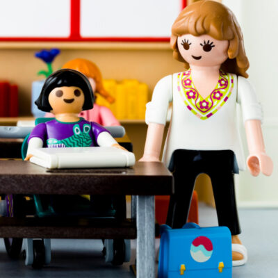 Unterrichtsszene mit einer Lehrerin, die bei einer Schülerin mit Arbeitslaptop steht mit Playmobil nachgestellt.