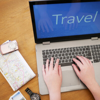 Ein Arbeitsplatz mit einem geöffneten Laptop, der das Wort „Travel“ auf dem Bildschirm zeigt. Auf dem Tisch liegen neben dem Laptop eine Landkarte, eine Kamera, ein paar Fotos und eine Armbanduhr. Die Person tippt auf der Tastatur, was darauf hinweist, dass sie eine Reise plant oder Informationen über Reiseziele recherchiert.