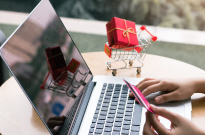 Symbolbild für PSN Gutscheinkarten, Person sitzt an LAptop und hält eine Bezahlkarte in der Hand, im Hintergrund ein Einkaufswagen mit einem verpackten GEschenk darin