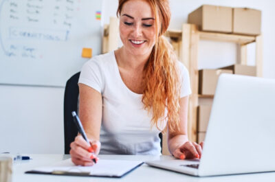 Eine junge Geschäftsinhaberin sitzt im Büro und schreibt Verkaufs- und Lieferdaten auf Papier, während sie einen Laptop benutzt.