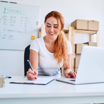 Eine junge Geschäftsinhaberin sitzt im Büro und schreibt Verkaufs- und Lieferdaten auf Papier, während sie einen Laptop benutzt.