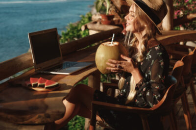 Eine junge Frau sitzt auf einer Holzterrasse mit Blick auf das Meer. Sie trägt einen Strohhut und ein florales Kleid und hält eine Kokosnuss mit Strohhalm in der Hand. Vor ihr auf dem Tisch steht ein Laptop und eine Platte mit Wassermelonenstücken. Sie scheint von einem Urlaubsort aus entspannt zu arbeiten.