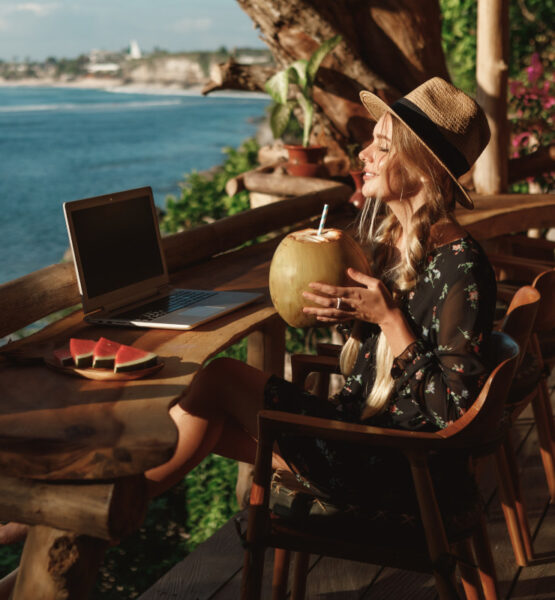 Eine junge Frau sitzt auf einer Holzterrasse mit Blick auf das Meer. Sie trägt einen Strohhut und ein florales Kleid und hält eine Kokosnuss mit Strohhalm in der Hand. Vor ihr auf dem Tisch steht ein Laptop und eine Platte mit Wassermelonenstücken. Sie scheint von einem Urlaubsort aus entspannt zu arbeiten.