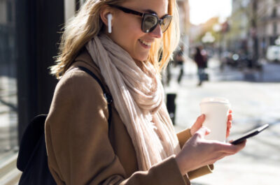 Eine junge Frau steht draußen in einer städtischen Umgebung und lächelt, während sie auf ihr Smartphone schaut. Sie trägt Sonnenbrillen, kabellose Ohrhörer, einen beigen Mantel und einen hellen Schal. In der linken Hand hält sie einen Kaffeebecher. Im Hintergrund sind verschwommen Gebäude und Fußgänger zu sehen, die eine belebte Straßenszene andeuten. Die Frau wirkt zufrieden und beschäftigt sich vermutlich mit einer Nachricht oder einem Anruf auf ihrem Smartphone.