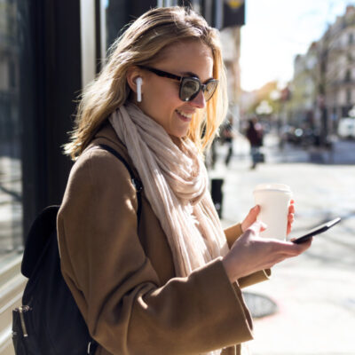 Eine junge Frau steht draußen in einer städtischen Umgebung und lächelt, während sie auf ihr Smartphone schaut. Sie trägt Sonnenbrillen, kabellose Ohrhörer, einen beigen Mantel und einen hellen Schal. In der linken Hand hält sie einen Kaffeebecher. Im Hintergrund sind verschwommen Gebäude und Fußgänger zu sehen, die eine belebte Straßenszene andeuten. Die Frau wirkt zufrieden und beschäftigt sich vermutlich mit einer Nachricht oder einem Anruf auf ihrem Smartphone.