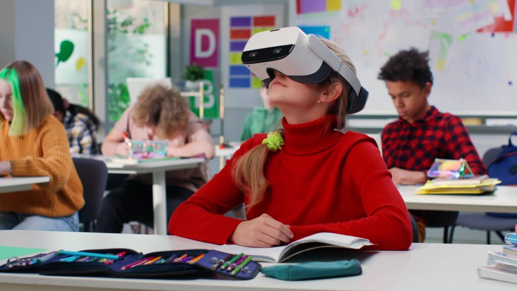 Ein junges Mädchen sitzt in einem Klassenzimmer und trägt ein Virtual-Reality-Headset. Sie hat einen roten Pullover an und schaut nach oben, während sie das Headset benutzt. Um sie herum sitzen andere Schüler, die konzentriert an ihren Aufgaben arbeiten.