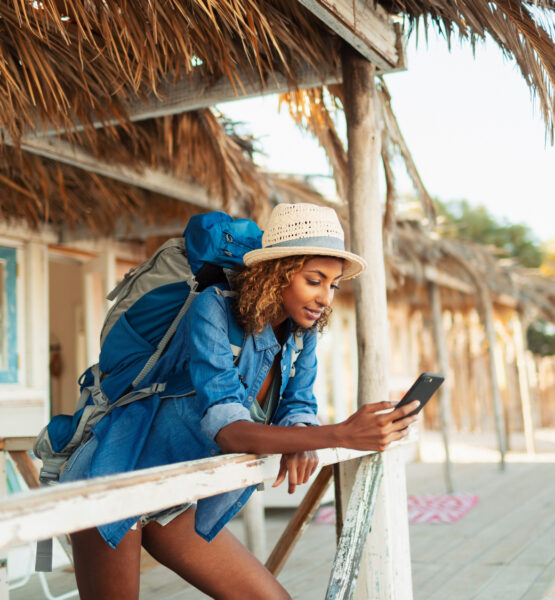 Das Bild zeigt eine junge Frau mit einem Rucksack, die sich an einem Holzgeländer lehnt. Sie trägt ein Strohhut und blickt konzentriert auf ihr Smartphone. Im Hintergrund ist eine rustikale, strandnahe Hütte mit einem Strohdach zu sehen. Die Szene wirkt sommerlich und entspannt, und die Frau scheint sich auf einer Reise zu befinden. Die Umgebung vermittelt eine tropische Atmosphäre mit hellen Farben und natürlicher Umgebung.