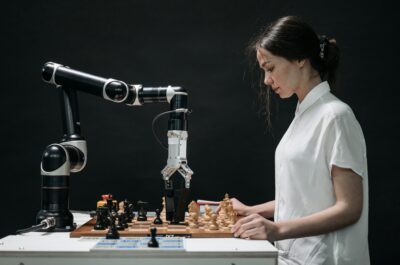 Eine Frau in einem weißen Hemd spielt Schach gegen einen Roboterarm. Der Roboter greift mit seiner mechanischen Hand nach einer Schachfigur auf einem Schachbrett. Die Szene symbolisiert den Wettstreit zwischen menschlicher Intuition und maschineller Präzision. Der Hintergrund ist dunkel, was die beiden Spieler hervorhebt.