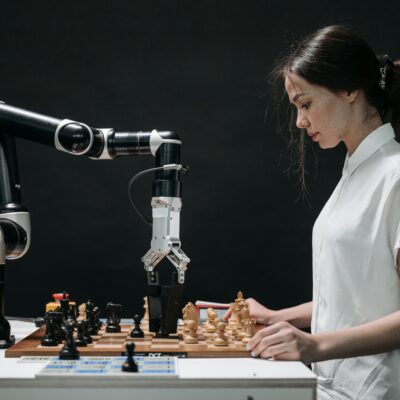 Eine Frau in einem weißen Hemd spielt Schach gegen einen Roboterarm. Der Roboter greift mit seiner mechanischen Hand nach einer Schachfigur auf einem Schachbrett. Die Szene symbolisiert den Wettstreit zwischen menschlicher Intuition und maschineller Präzision. Der Hintergrund ist dunkel, was die beiden Spieler hervorhebt.