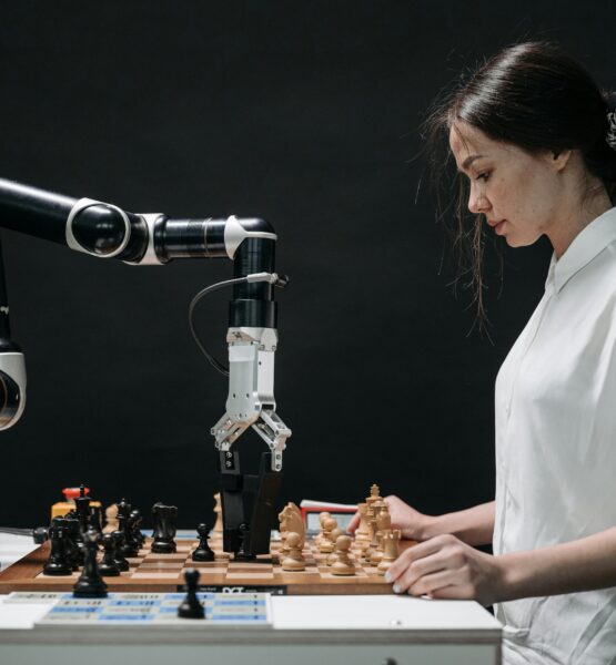 Eine Frau in einem weißen Hemd spielt Schach gegen einen Roboterarm. Der Roboter greift mit seiner mechanischen Hand nach einer Schachfigur auf einem Schachbrett. Die Szene symbolisiert den Wettstreit zwischen menschlicher Intuition und maschineller Präzision. Der Hintergrund ist dunkel, was die beiden Spieler hervorhebt.