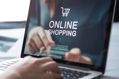 Eine Person nutzt einen Laptop für Online-Shopping, hält eine Kreditkarte in der Hand und tippt auf der Tastatur. Auf dem Bildschirm ist der Schriftzug 'Online Shopping' mit einem Einkaufswagensymbol zu sehen. Im Hintergrund befindet sich eine unscharfe, dunkle Umgebung.