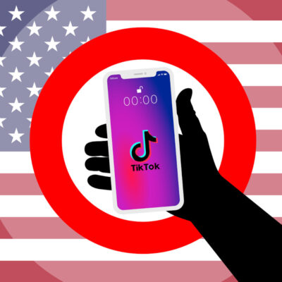 Ein Verbotsschild mit einem in der Hand gehaltenen Handy auf der TikTok läuft. Im Hintergrund ist die amerikanische Flagge zu sehen.