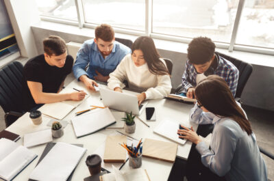 Eine Gruppe von Studenten arbeitet gemeinsam an einem Tisch mit Laptops. Sie sind in eine konzentrierte Teamarbeit vertieft und umgeben von Schreibmaterialien wie Stifte, Notizbücher und Ordner. Die Szene vermittelt ein produktives Lernumfeld mit moderner Technologie.